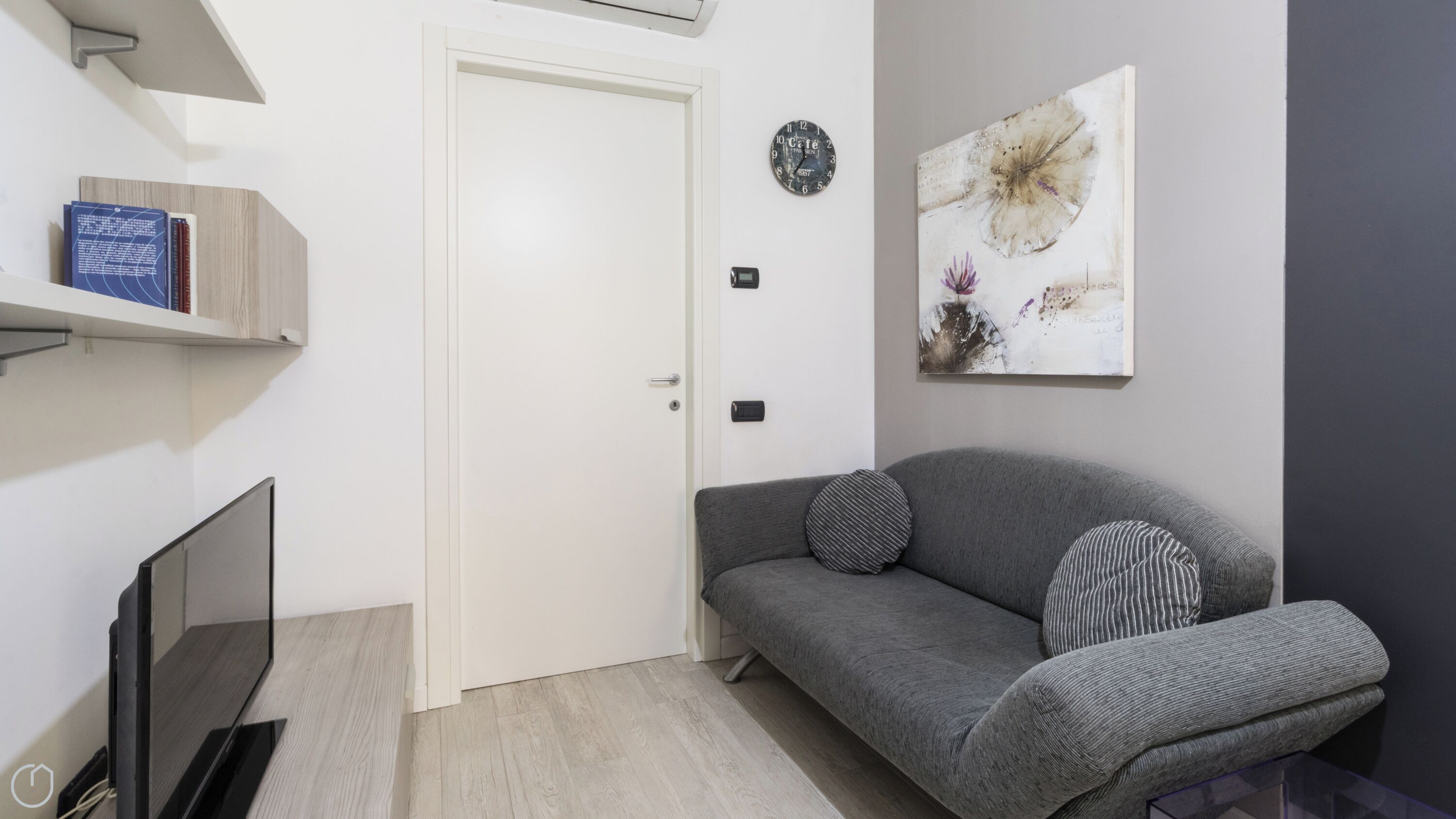 A modern living room with a grey sofa, a flat-screen TV, a white door, and a wall-mounted clock.