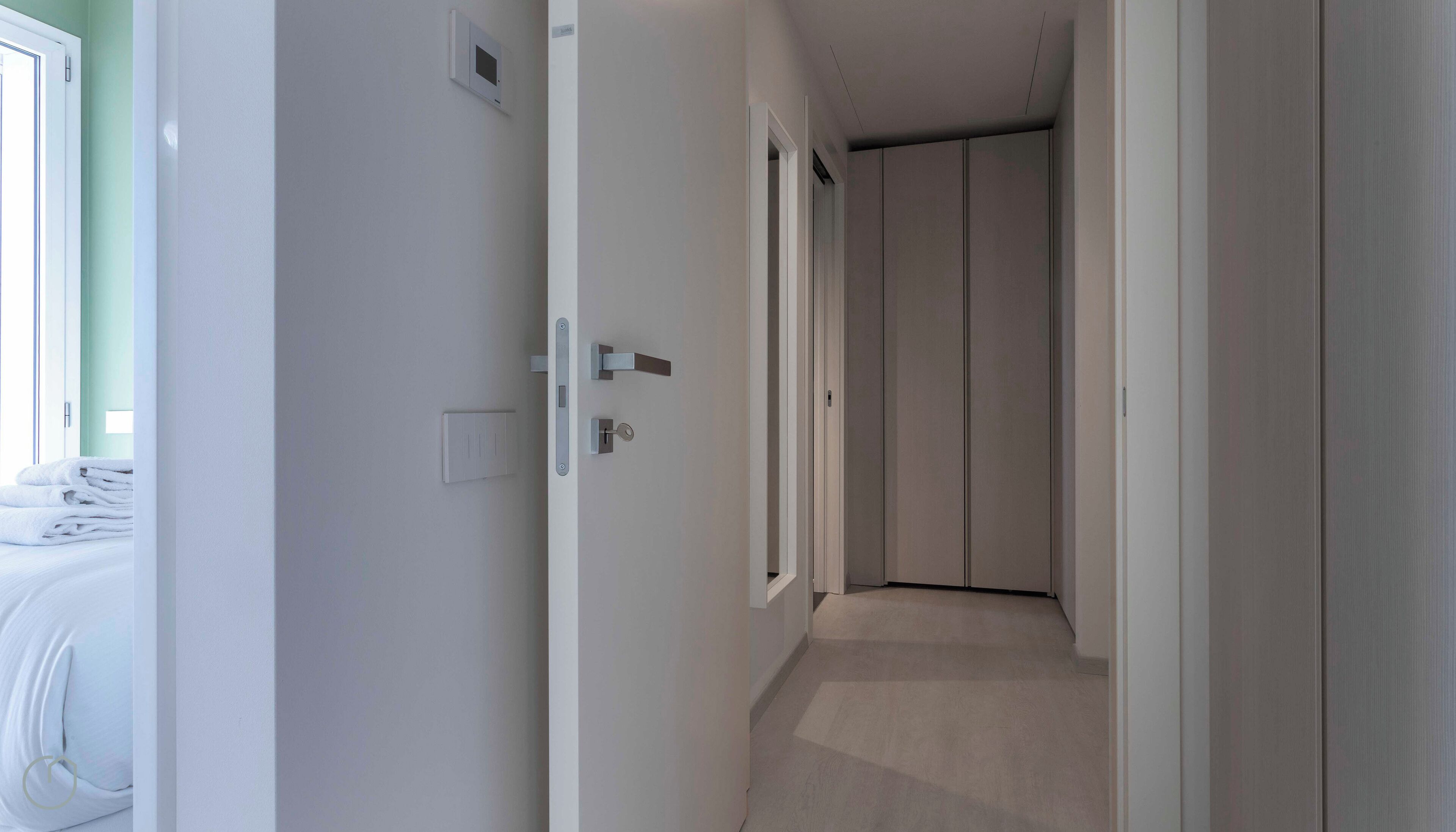 A narrow hallway with a carpeted floor, white walls, and a door leading to a room with a bed and a window.
