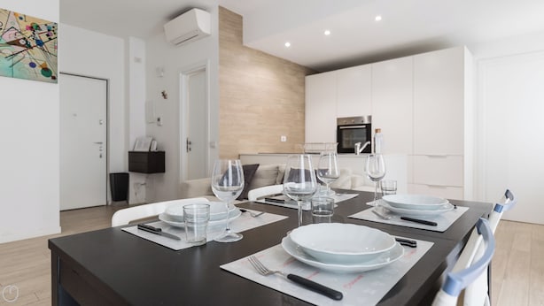 A modern dining area with a dark wooden table set for four, white plates, clear glasses, and cutlery. A kitchen with white cabinets and a built-in oven is visible in the background. - Italianway - Spontini 10 (Milan)