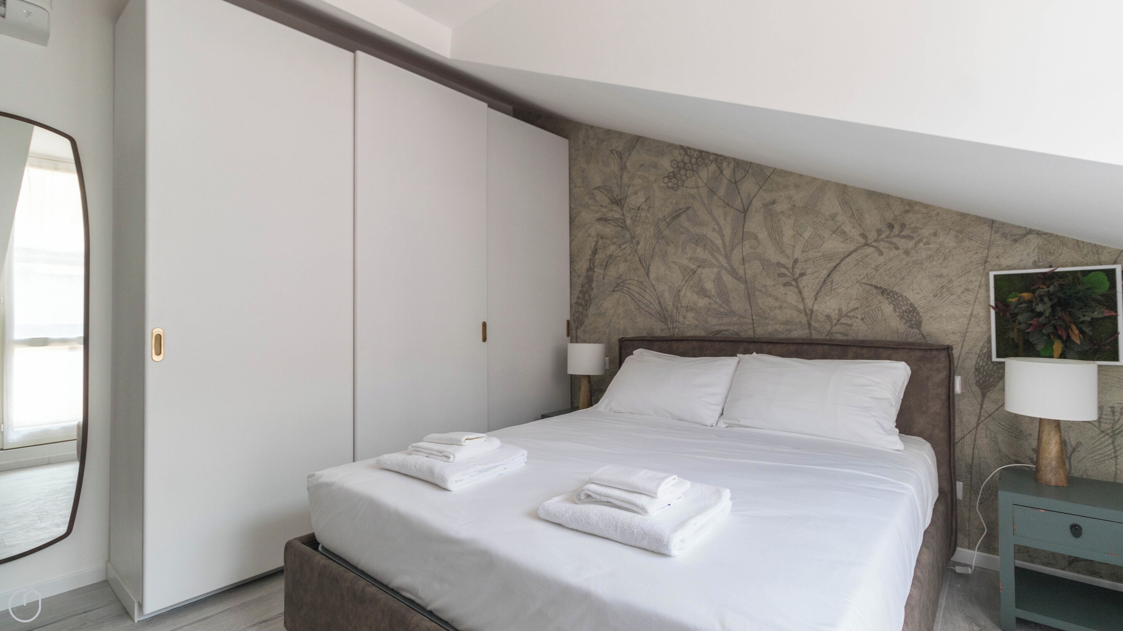 Een netjes opgemaakt bed met witte lakens en handdoeken, een houten hoofdbord, een nachtkastje met een lamp en een garderobe met schuifdeuren.