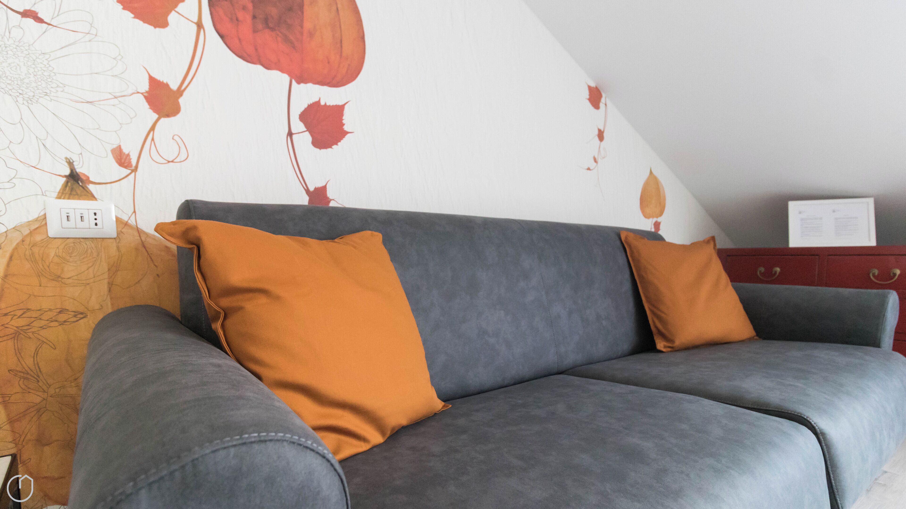 Un sofá gris con dos cojines naranjas, un armario rojo y una pared con diseño floral.