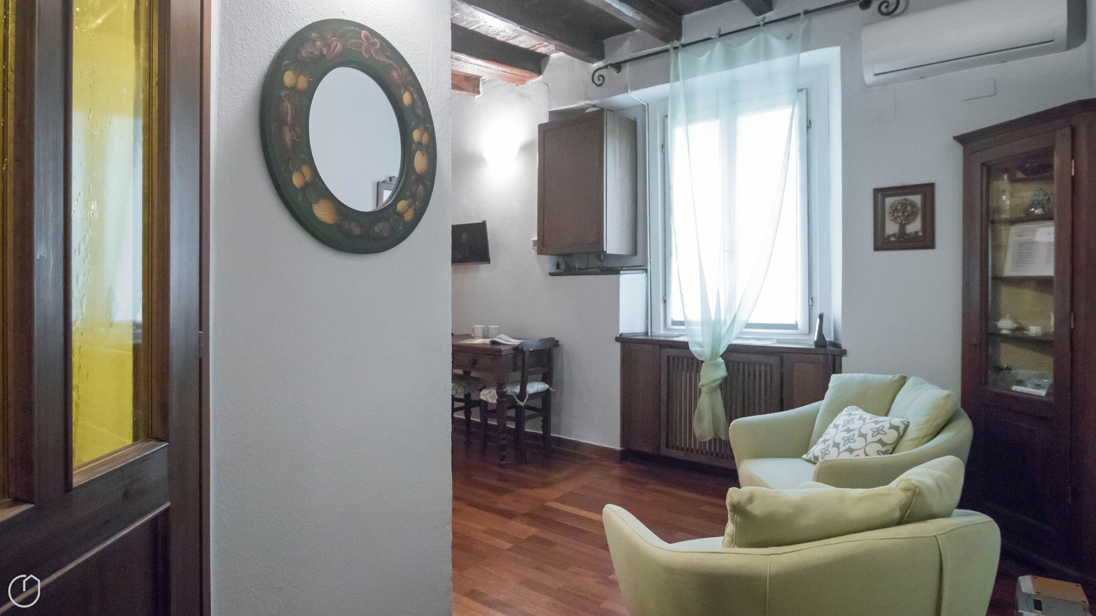 Un soggiorno con pavimento in legno, una poltrona verde, un tavolo da pranzo con sedie, uno specchio e una finestra con tende leggere.