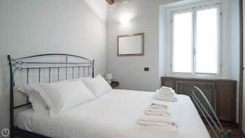 Un letto rifatto con lenzuola bianche e una testiera in metallo, una lampada sul comodino, una finestra con tende bianche e un termosifone sotto la finestra.
