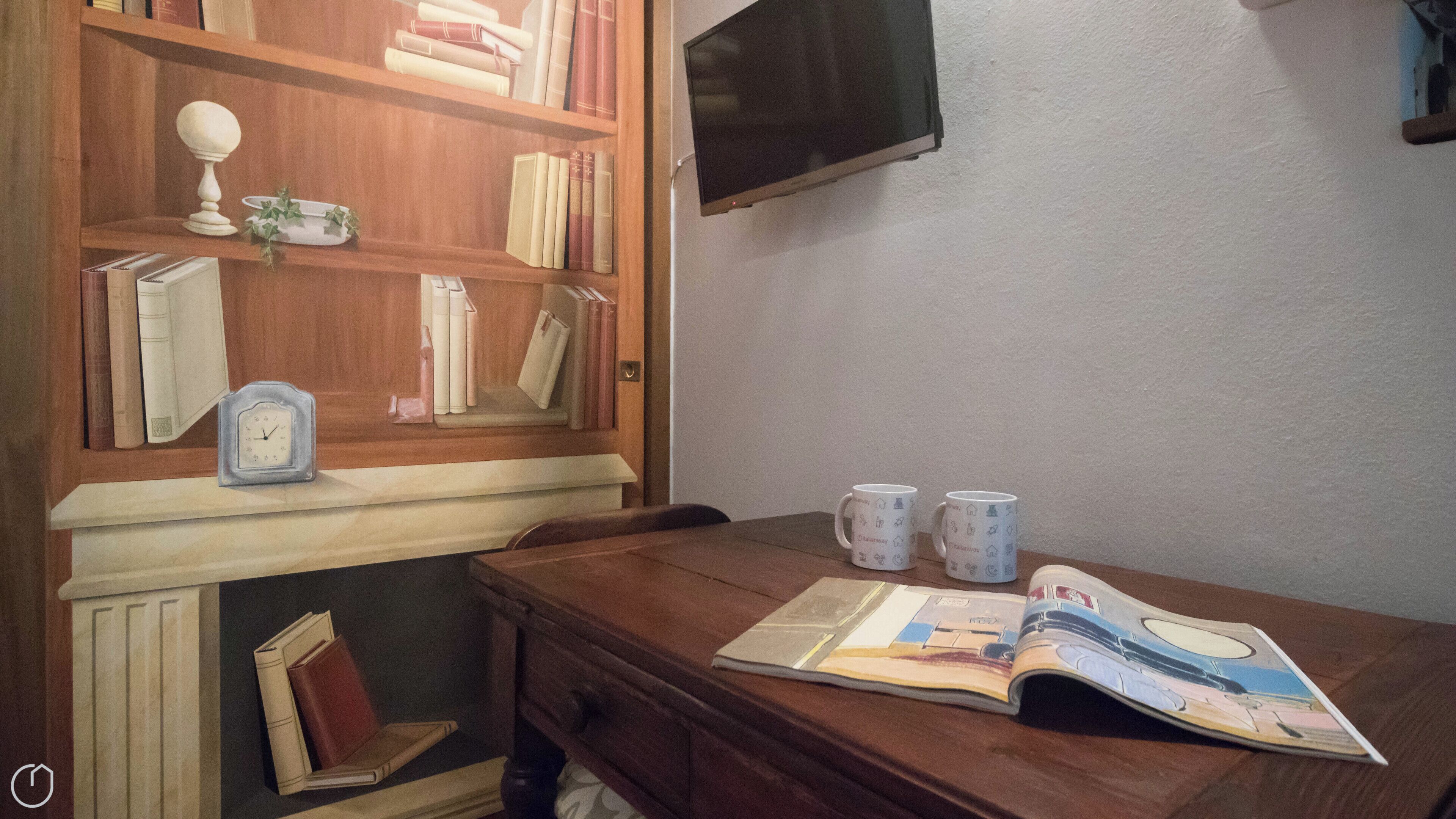 Una stanza con una libreria in legno, una televisione fissata al muro e un tavolo con una rivista e due tazze.