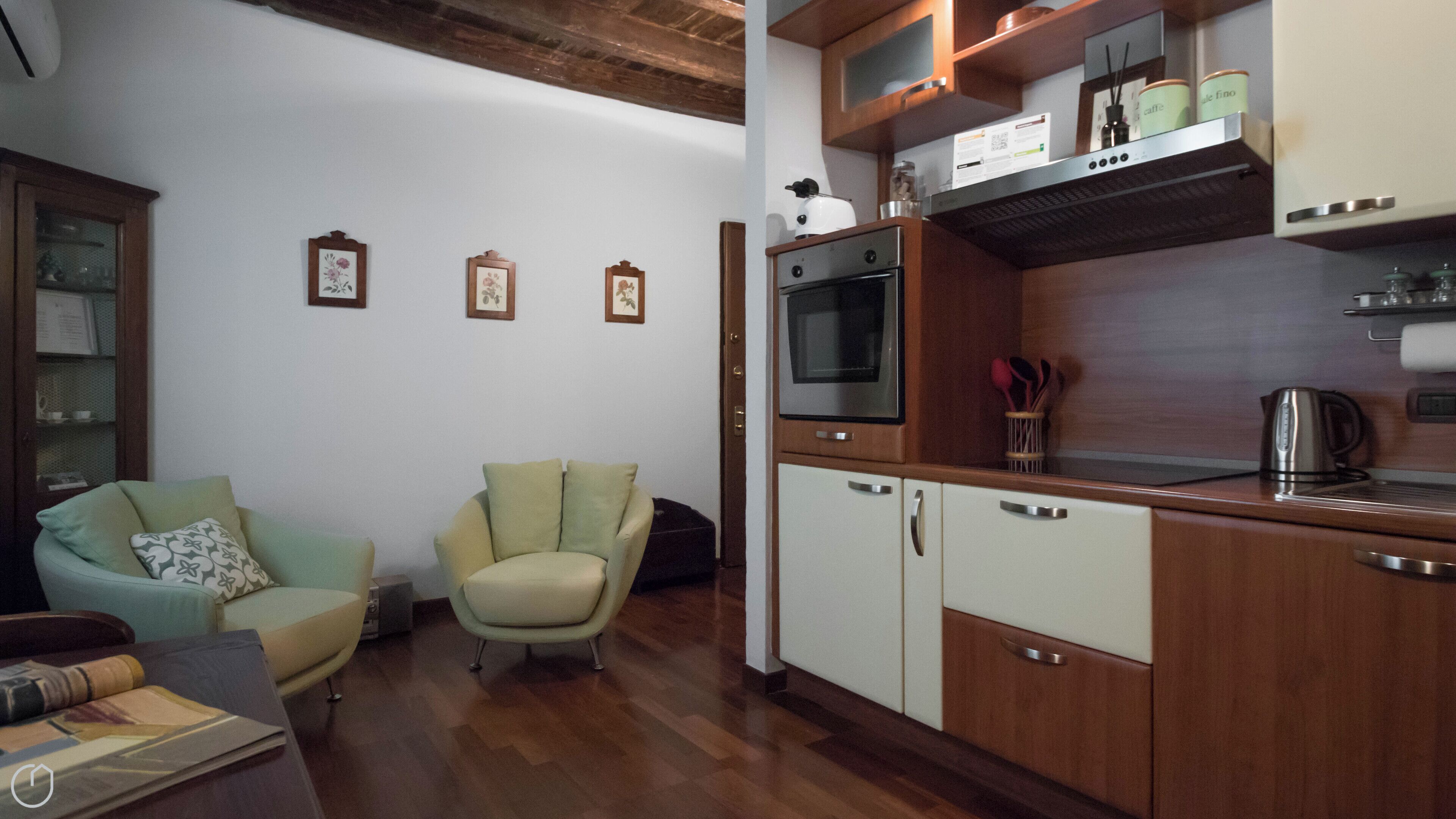 Una cucina compatta con mobili in legno, forno integrato e microonde. Si notano quadri appesi al muro e un piccolo angolo salotto con una poltrona e un divano.
