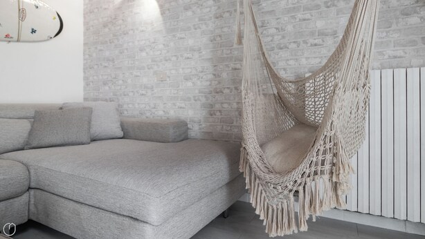 Ein modernes Wohnzimmer mit einem grauen Sofa, einem Hängesessel aus Makramee und einer Wand mit Ziegeloptik.