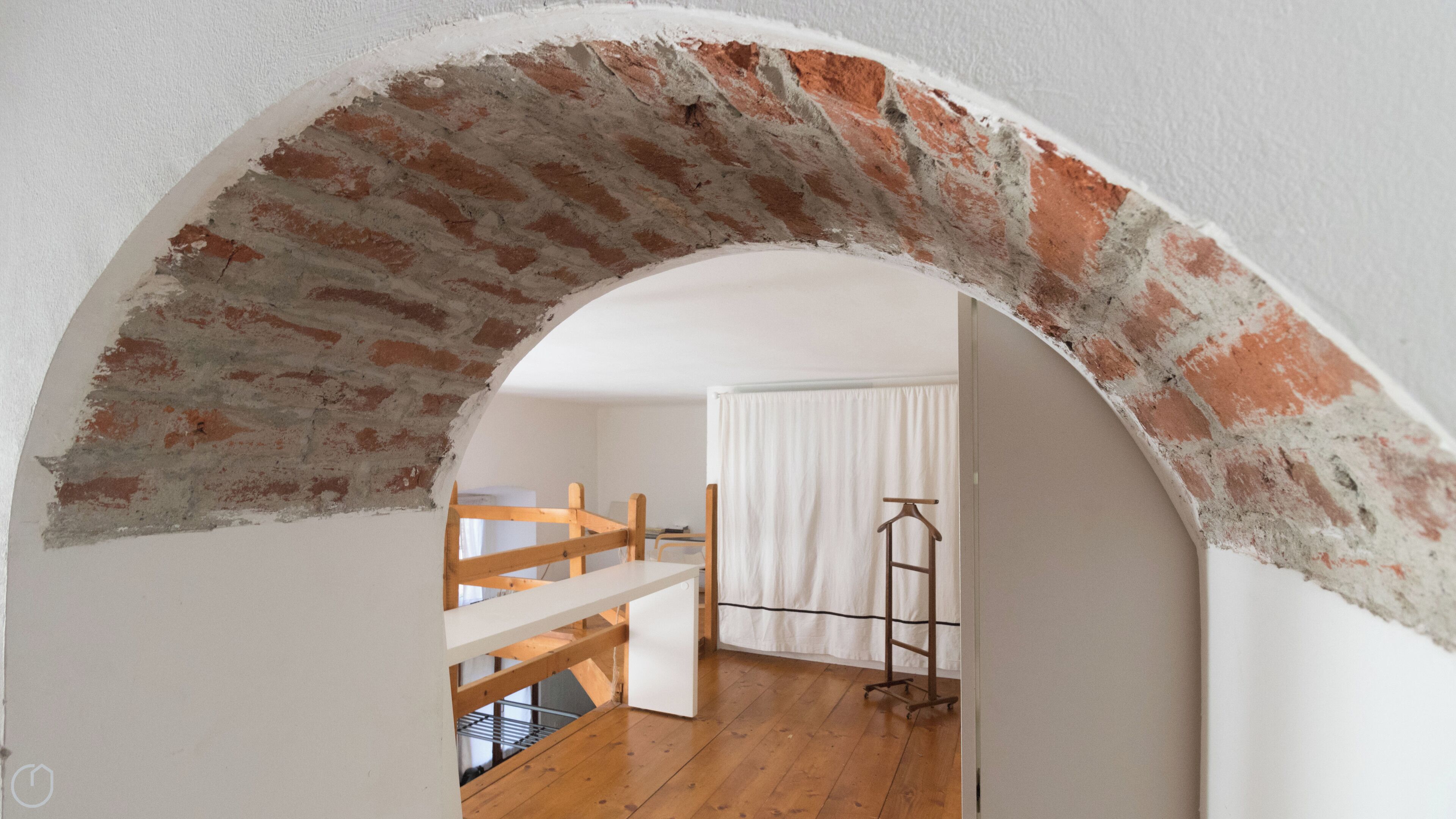 Un interior con un arco de ladrillo que conduce a una habitación con una cama, una silla de madera y un perchero.
