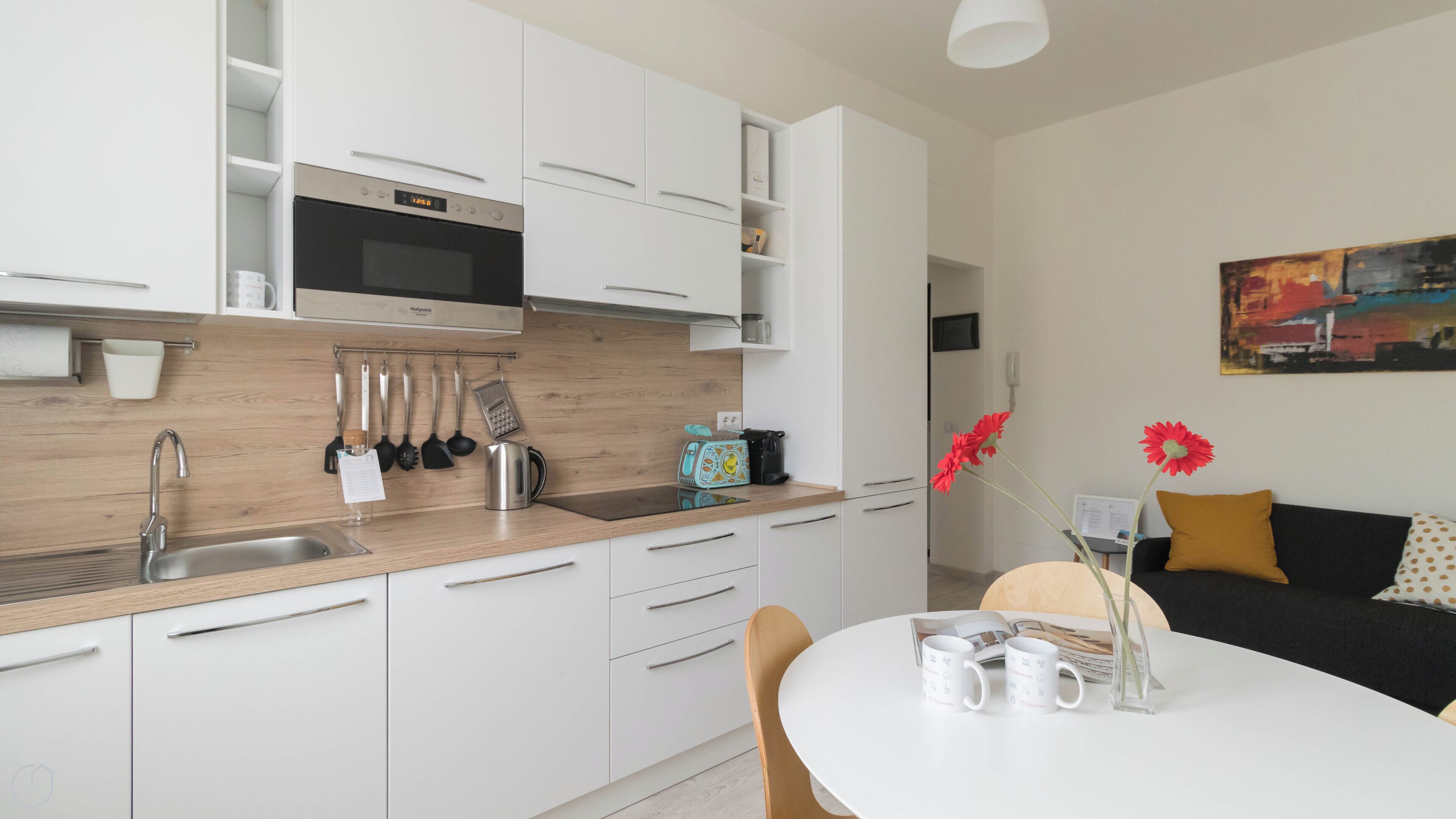 A modern kitchen with white cabinets, a stainless steel sink, and a microwave. There is a dining area with a white table and chairs, and a black sofa with yellow cushions.