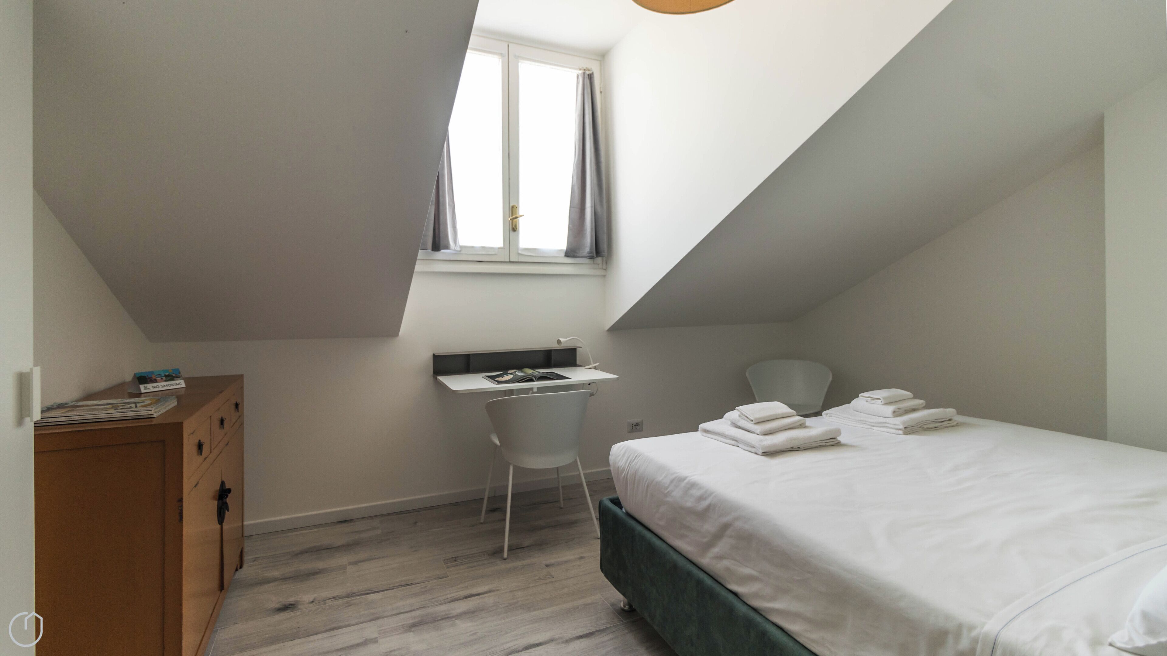 Un dormitorio pequeño y moderno con techo inclinado, un escritorio, una silla, una cama con toallas ordenadas y un aparador de madera.
