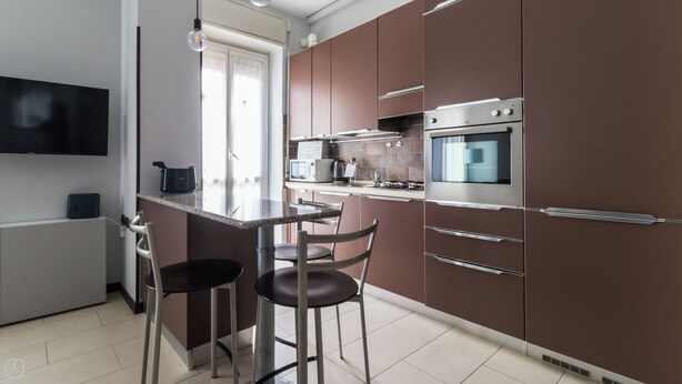 A modern kitchen with a flat-screen TV, a built-in oven, and a microwave. There are two bar stools and a dining table.