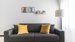 A grey sofa with two mustard-colored cushions, a black and white lamp, and two framed posters on the wall.