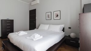 A modern bedroom with a large bed, a dark wardrobe, a bedside table with a lamp, and two framed pictures on the wall.