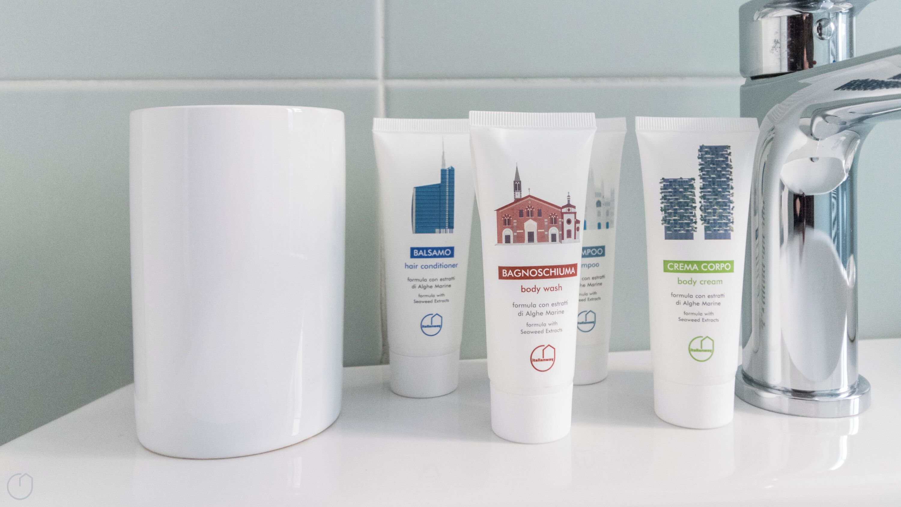 A bathroom with a white sink, a white mug, and three white bottles of toiletries labeled Balsamo, Bagnosciumia, and Crema Corpo.