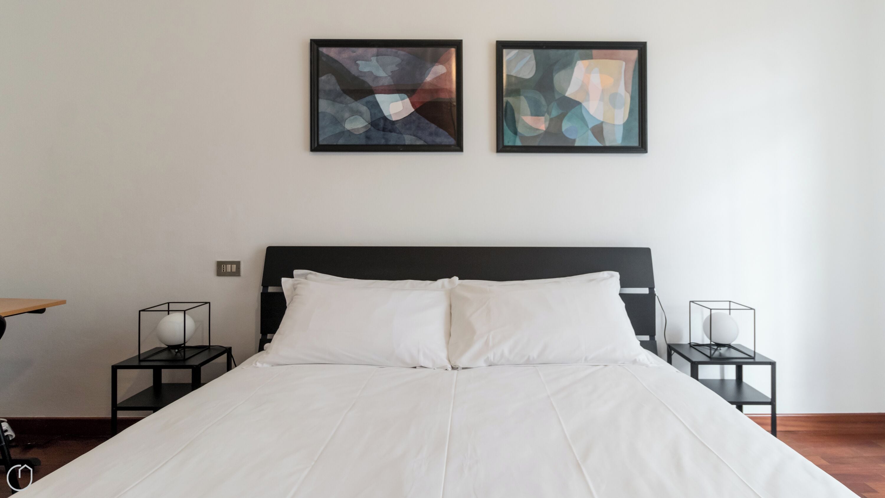 Una cama bien hecha con sábanas blancas, dos cuadros abstractos en la pared y un suelo de madera.
