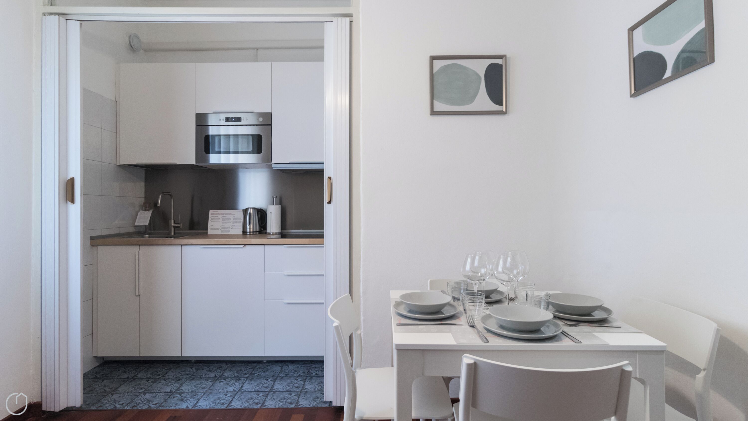 Cozinha compacta com forno embutido, cafeteira e mesa de jantar posta para quatro, com pratos, copos e talheres.