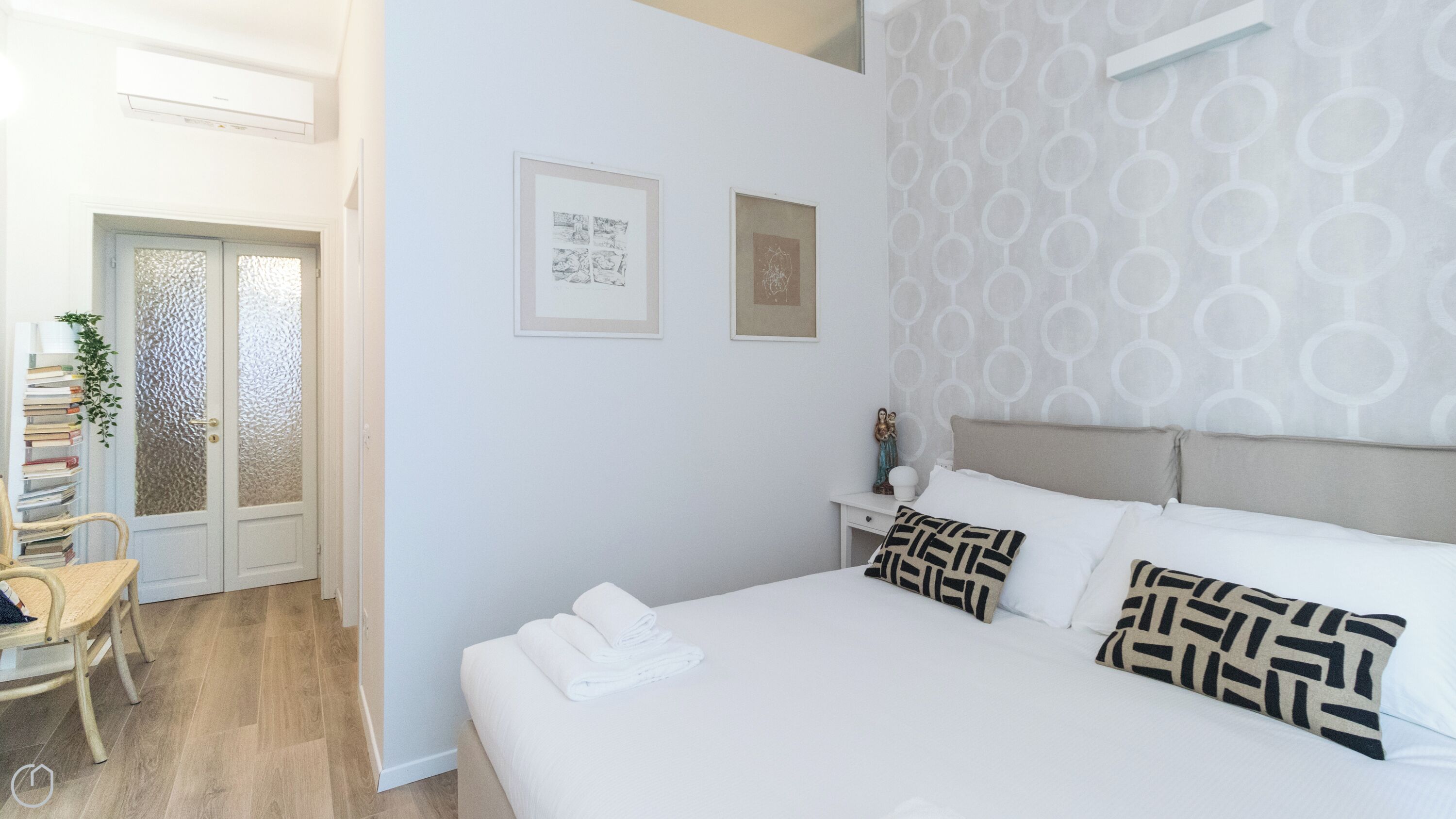 A bedroom with a large bed, two pillows, a nightstand, a wall with two framed pictures, a door with frosted glass, and a chair with books.