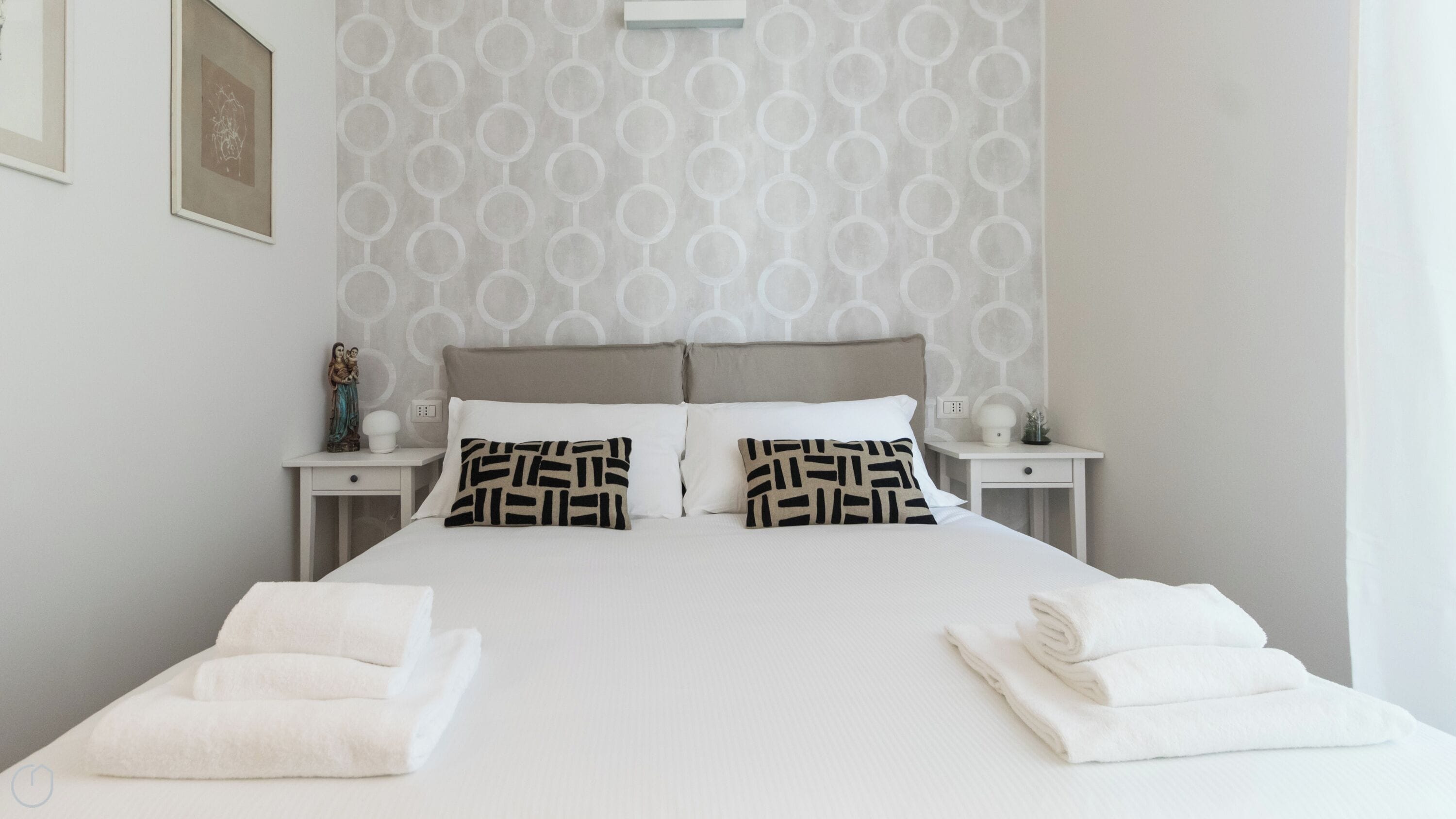 A neatly made bed with white linens and patterned pillows, two bedside tables with decorative items, and a wall with a circular pattern.