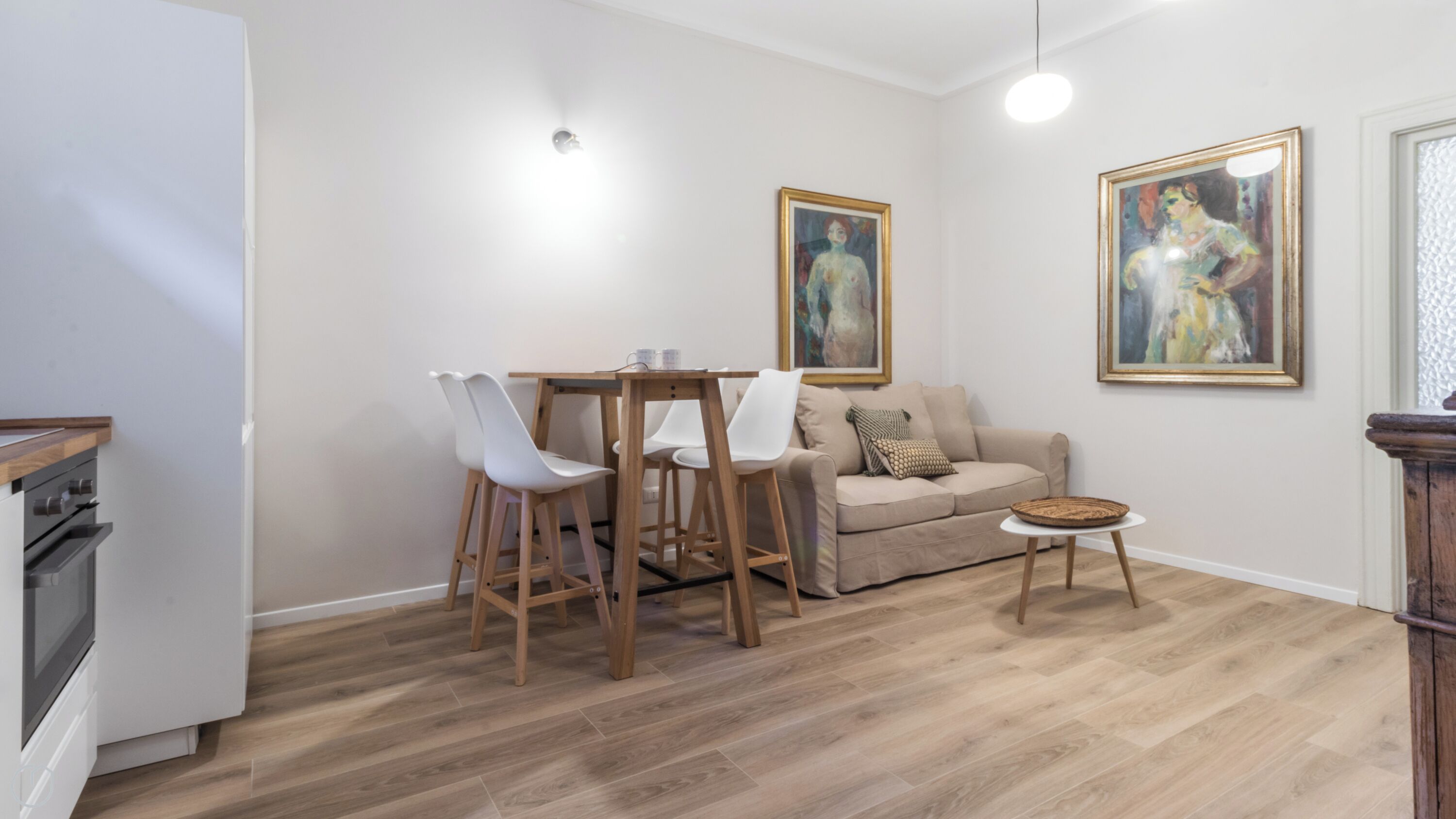 A modern living room with a wooden dining table, white chairs, a beige sofa, and two framed paintings on the wall.