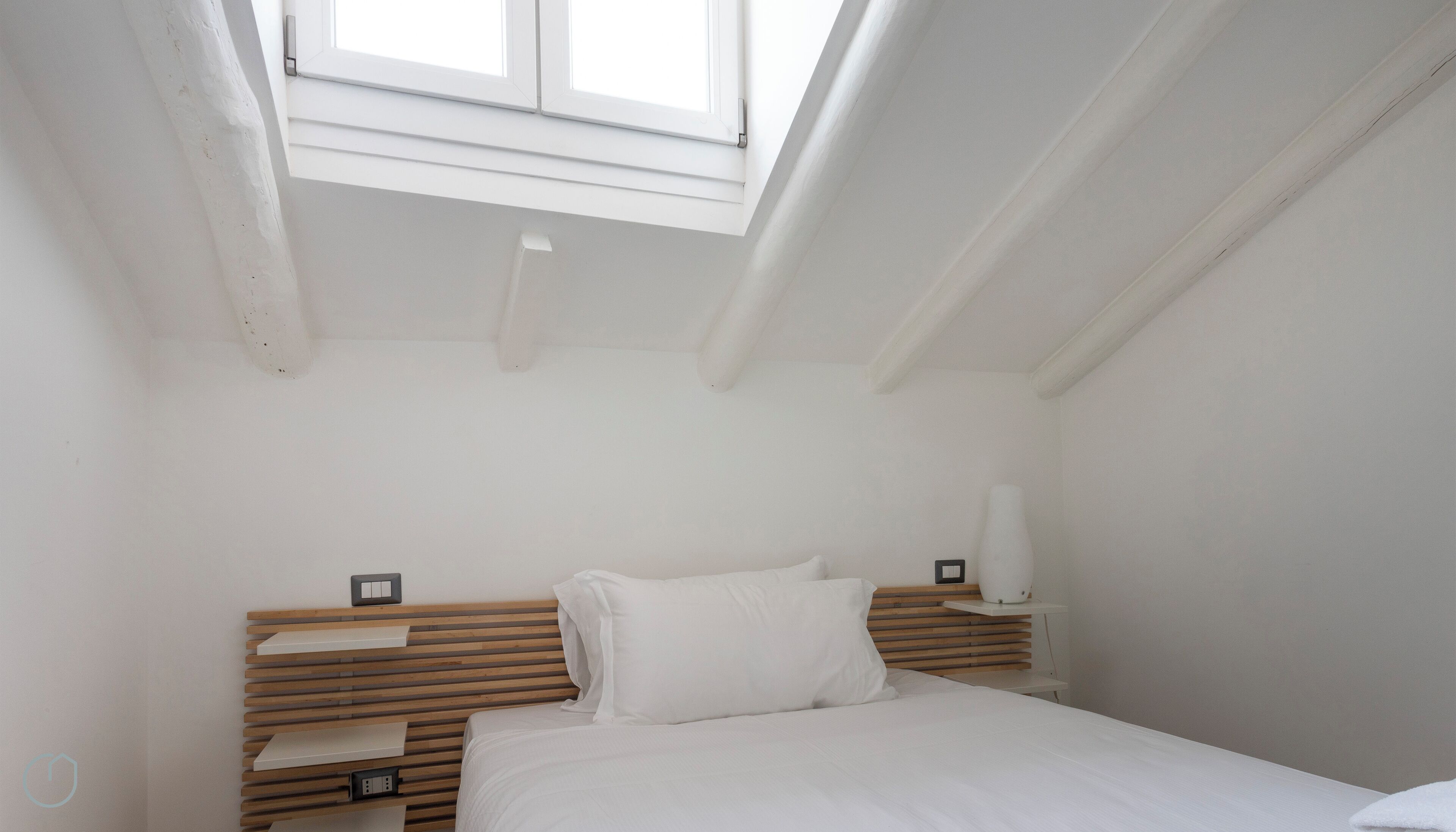 Ein Schlafzimmer mit Dachfenster, einem Holz-Kopfteil, einem Bett mit weißer Bettwäsche und einem Nachttisch mit Lampe.
