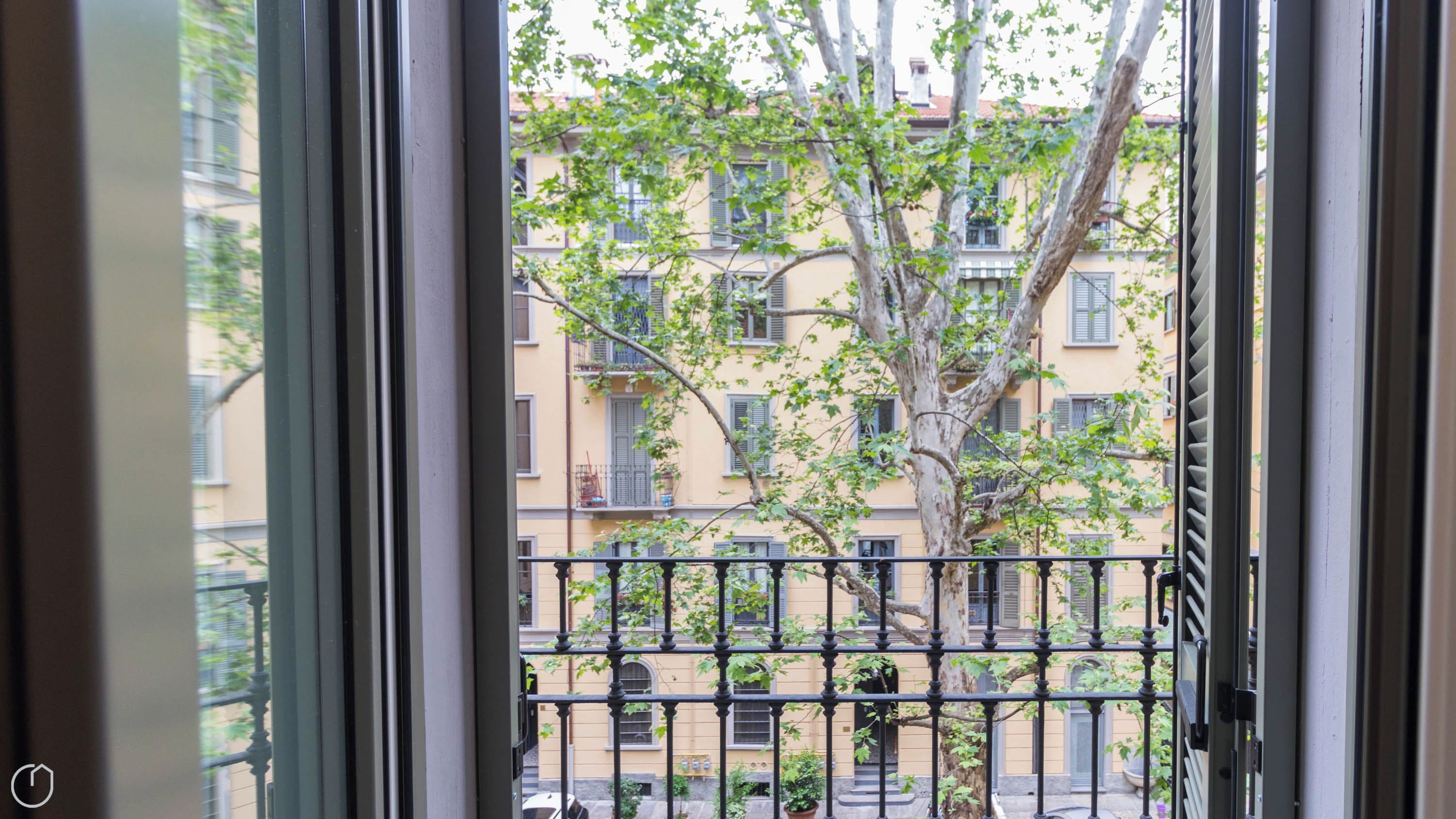 A view through a window with a metal railing, a tree, and a building with multiple windows and balconies.