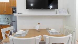 Ein Essbereich mit Holztisch, gedeckt für zwei Personen, weißen Stühlen, einem über dem Tisch montierten Fernseher und einer Küchenzeile mit Kühlschrank und Mikrowelle links.