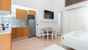 Eine kompakte Küche mit Holzausbau, einem Kühlschrank und einer Mikrowelle. Ein Esstisch mit Stühlen sowie ein weißes Bett mit Handtüchern.