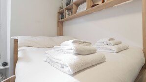 En pænt redt seng med hvide sengetøj og håndklæder, et træhovedgærde og en hylde med bøger og en lampe.