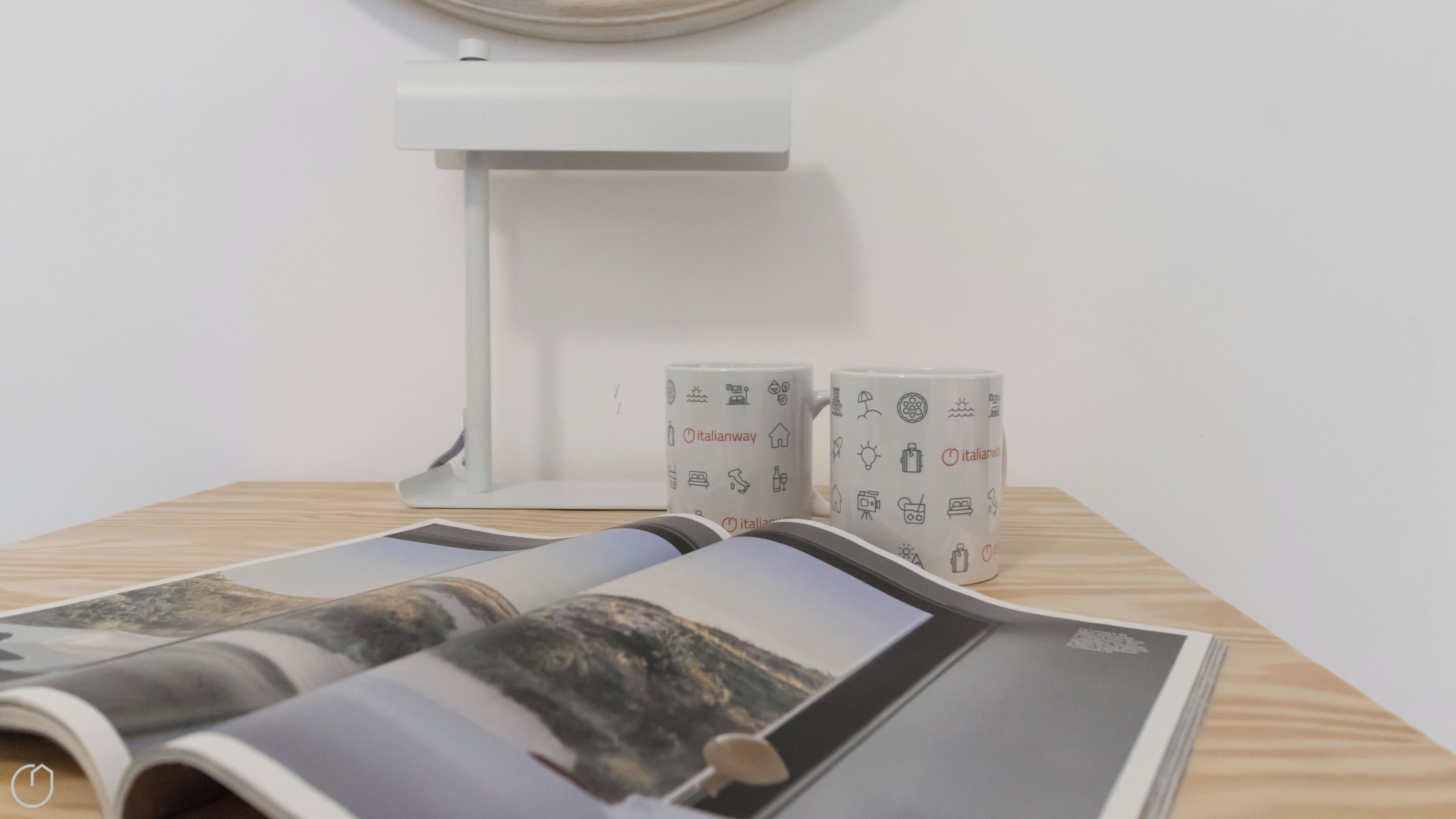 Une table avec un magazine, deux tasses ornées du logo « Italianway » et une lampe de bureau blanche.