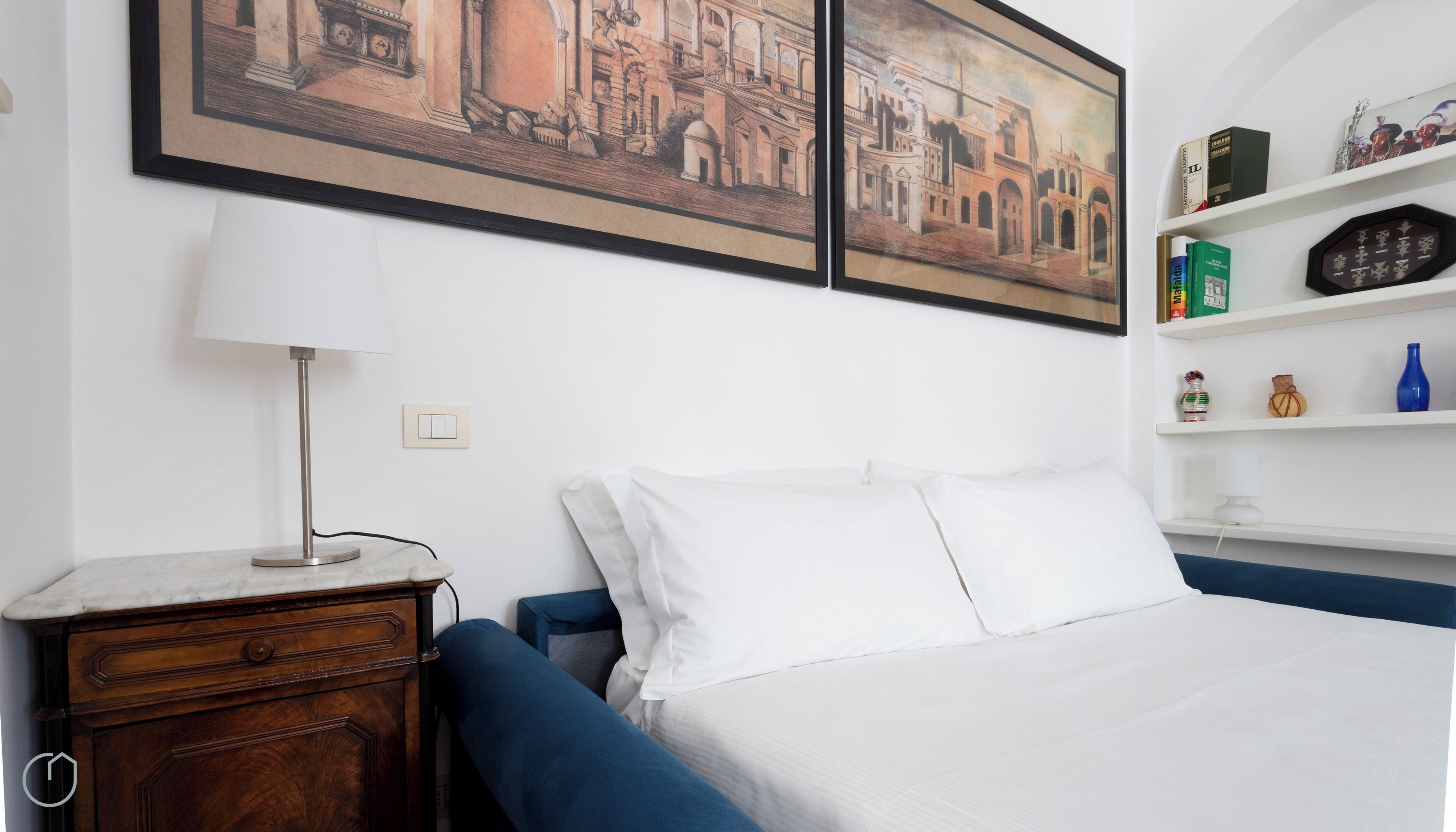 A bedroom with a bed, a nightstand, a lamp, a dresser, and a shelf with books and decorative items.