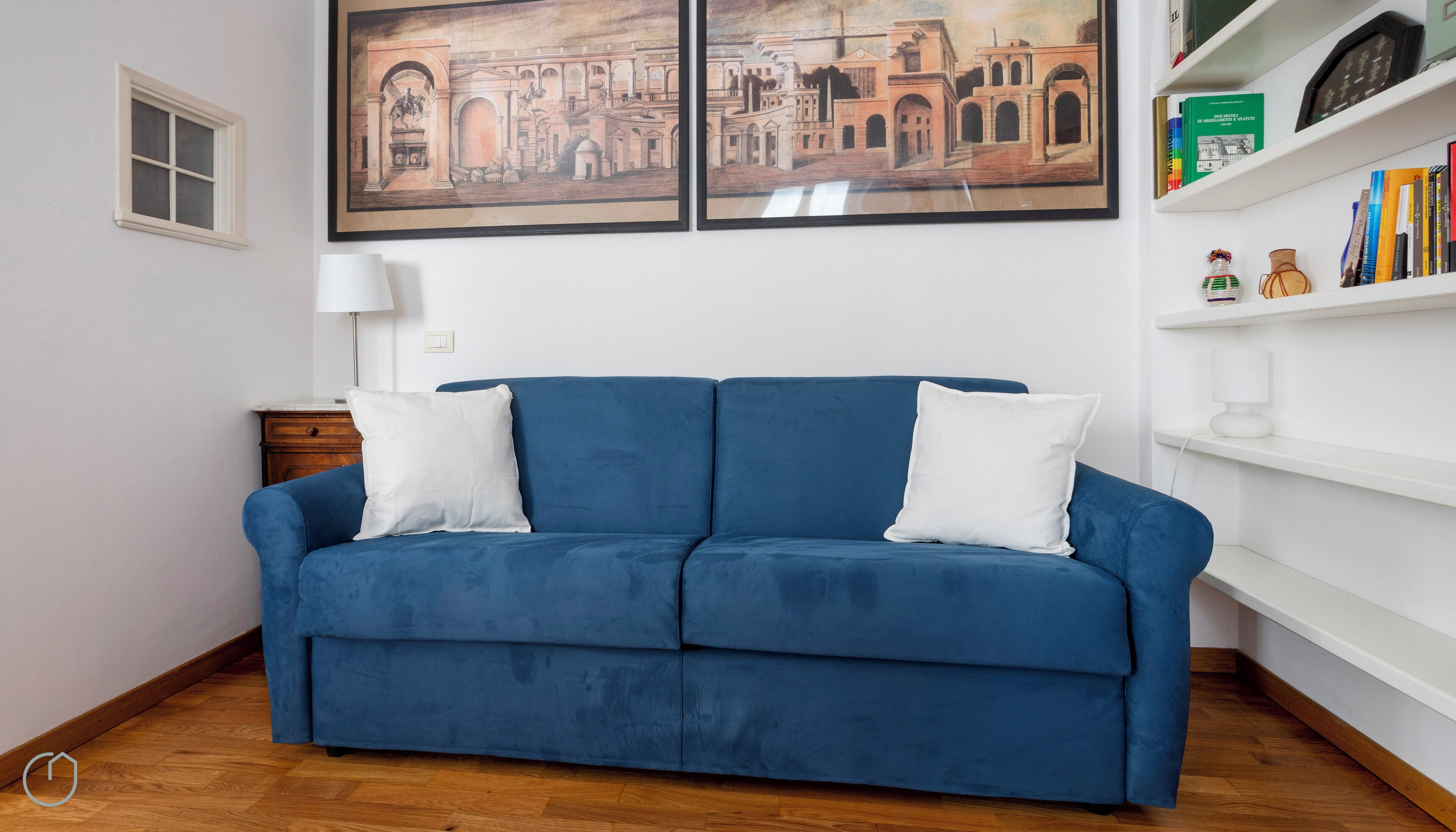 Un canapé bleu avec des coussins blancs, un plancher en bois, un mur blanc, une petite fenêtre et deux tableaux encadrés au-dessus du canapé.