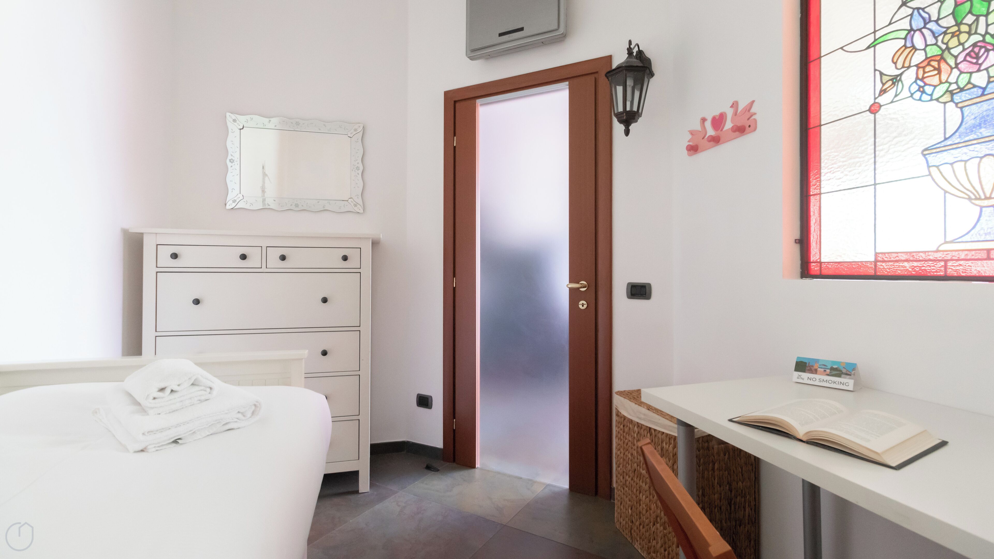 A bedroom with a bed, a dresser, a door, a wall-mounted air conditioner, a wall-mounted light fixture, a stained glass window, and a 'No Smoking' sign.
