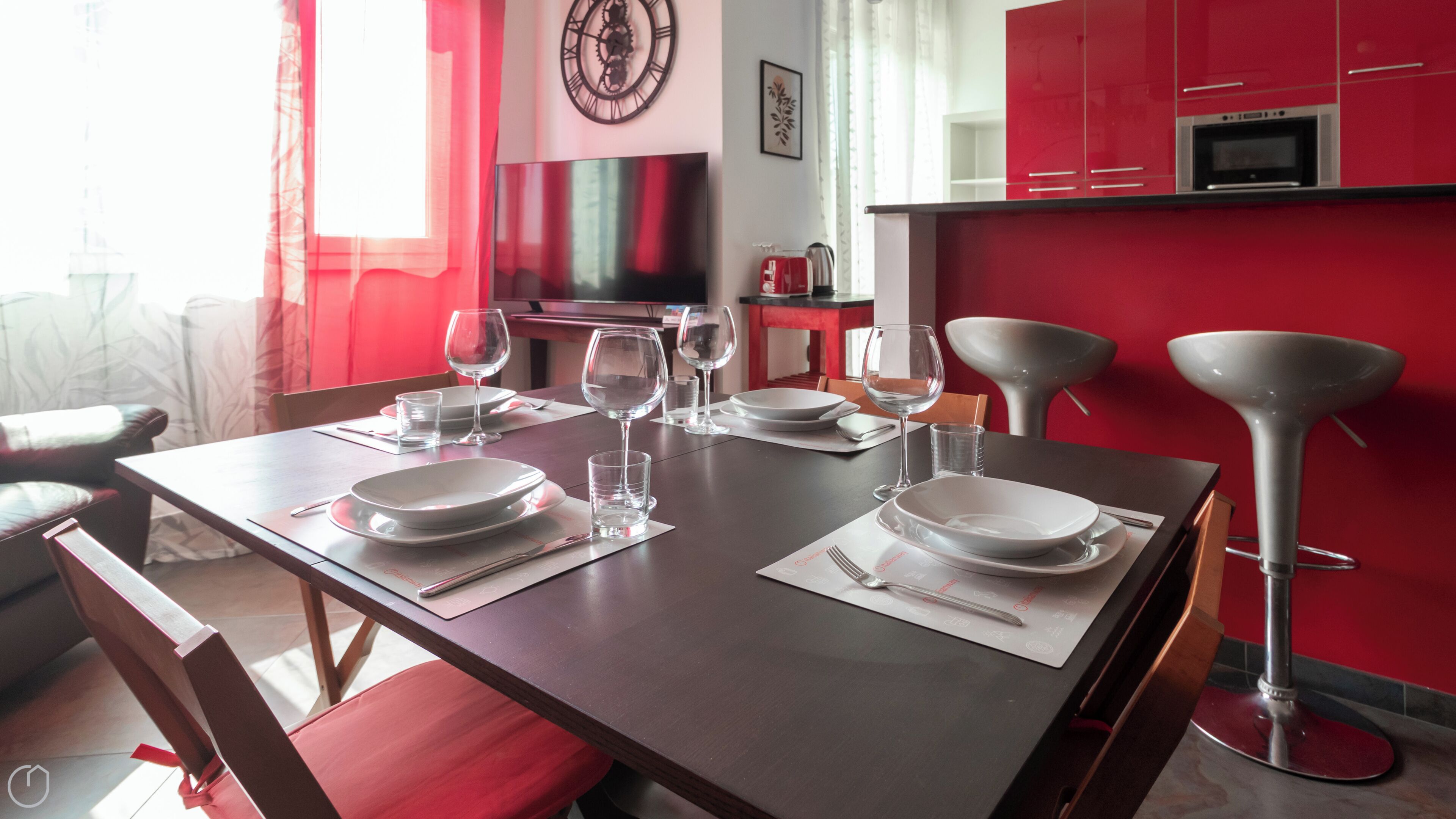 Une table à manger dressée pour quatre personnes, avec assiettes, verres et couverts, une cuisine rouge en arrière-plan et une lampe moderne.