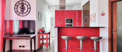 A modern kitchen with red cabinets, a built-in oven, and a microwave. There is a dining area with a TV, a red table, and three bar stools.