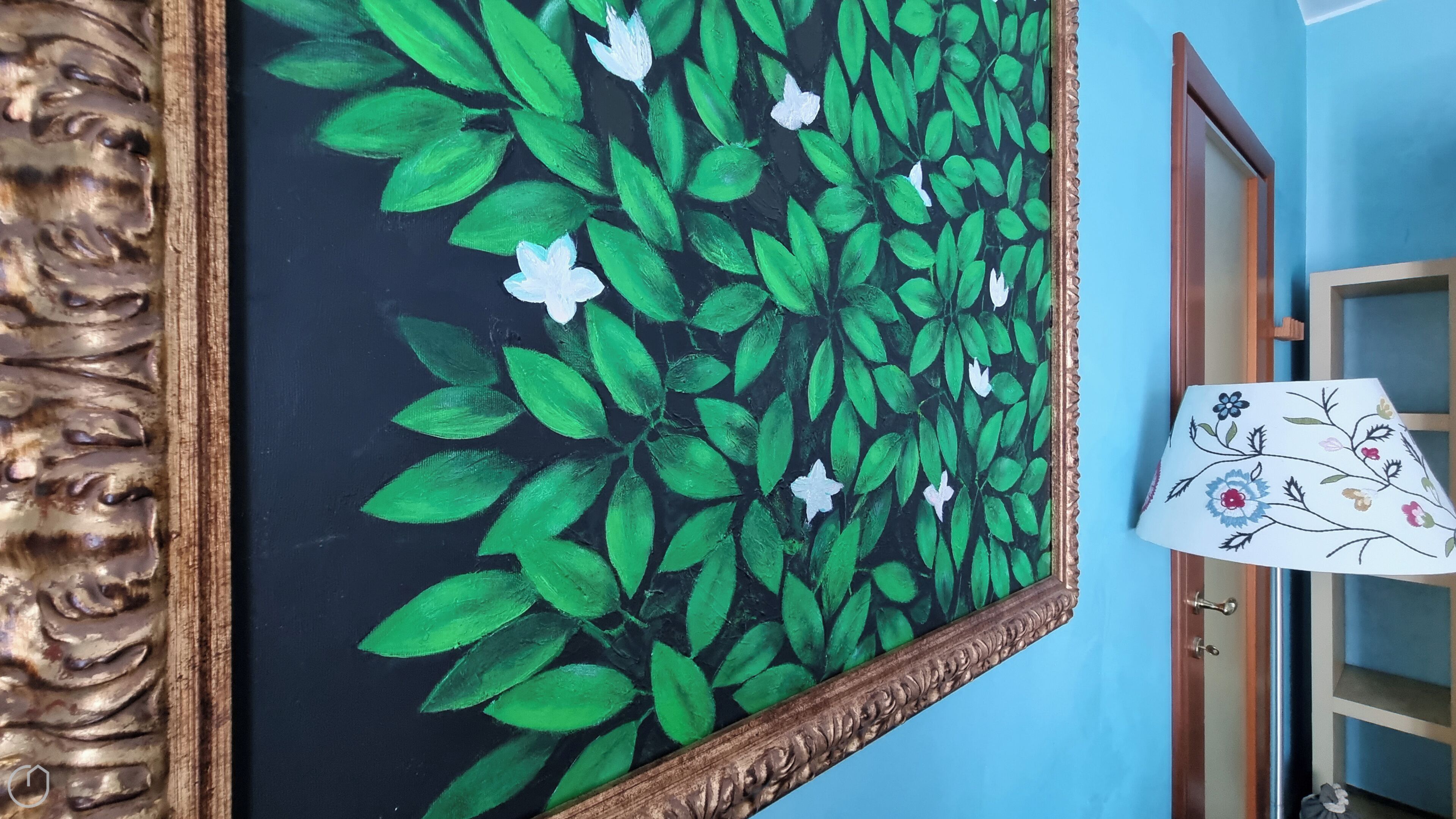 A painting of green leaves and white flowers on a black background, framed in gold, hanging on a blue wall.