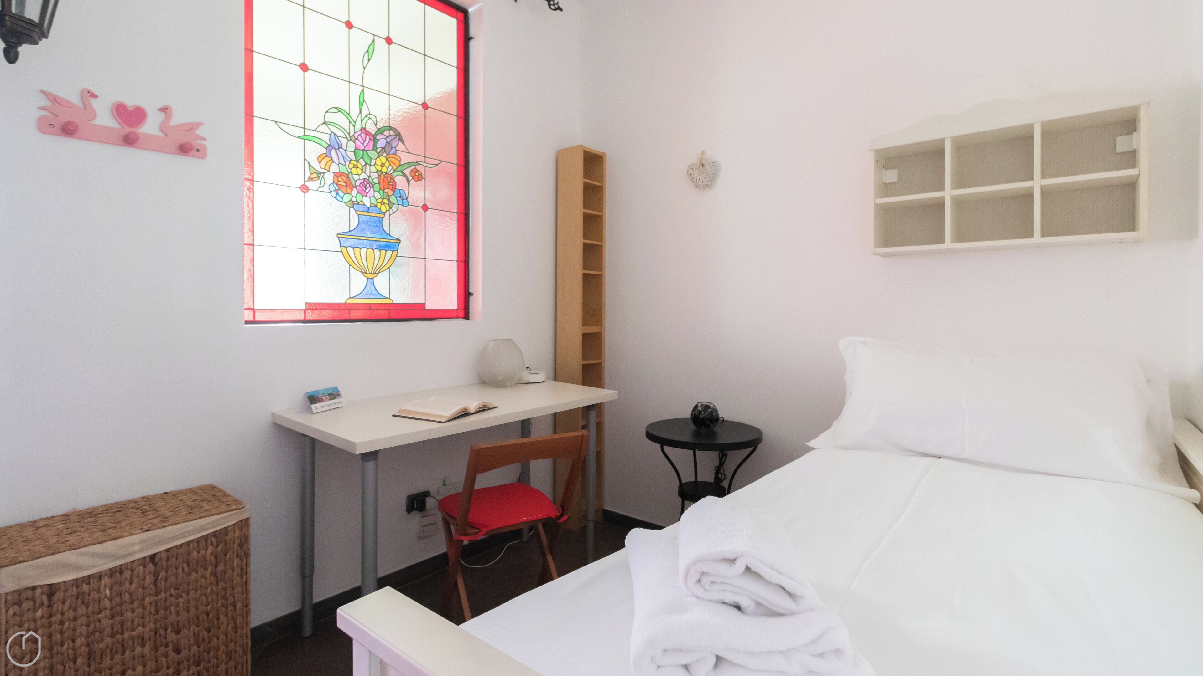 A bedroom with a bed, a desk with a chair, a small table, a bookshelf, and a stained glass window.