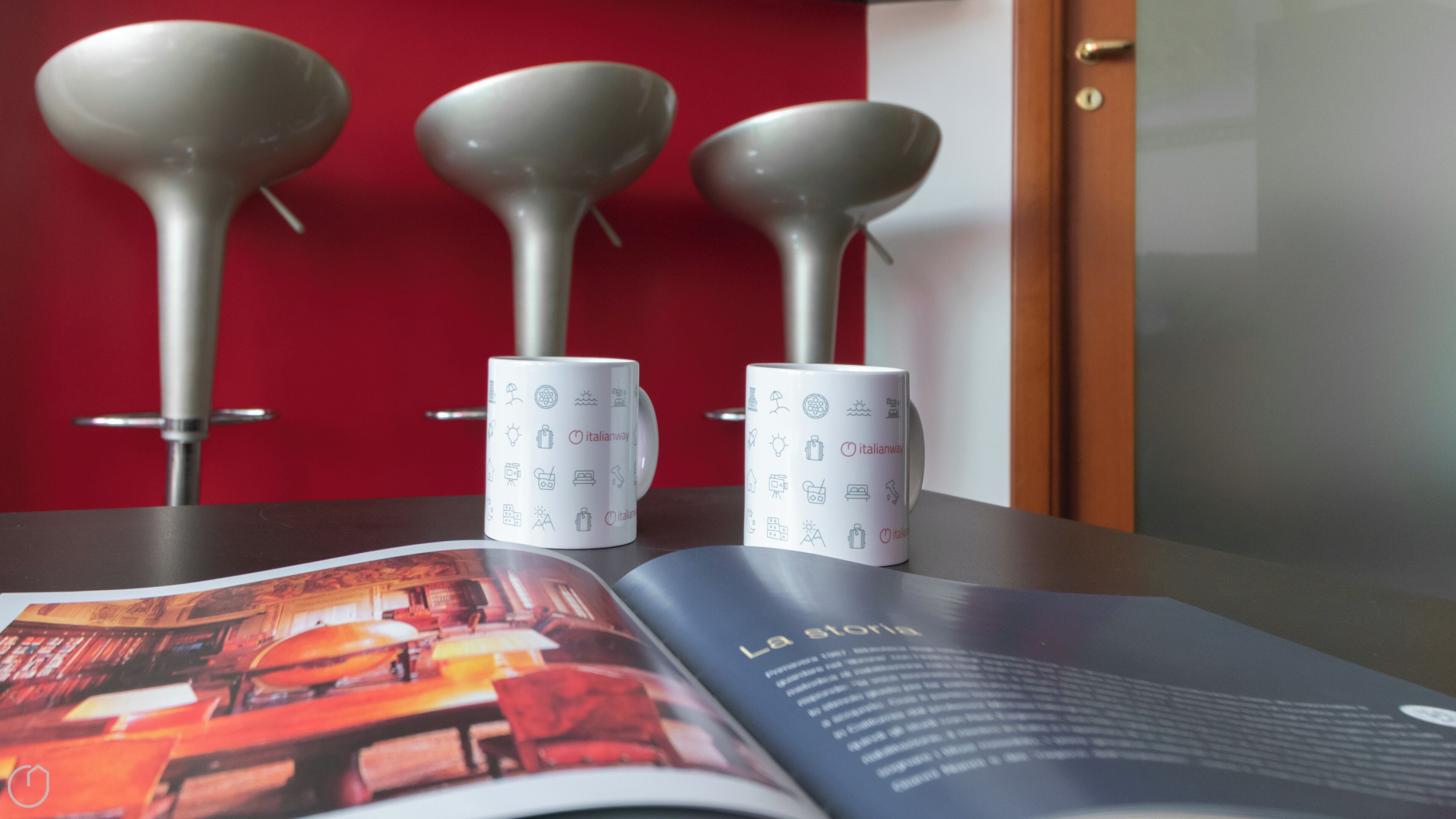 Three white mugs with Italian symbols on a table, a magazine with a red cover, and a door in the background.