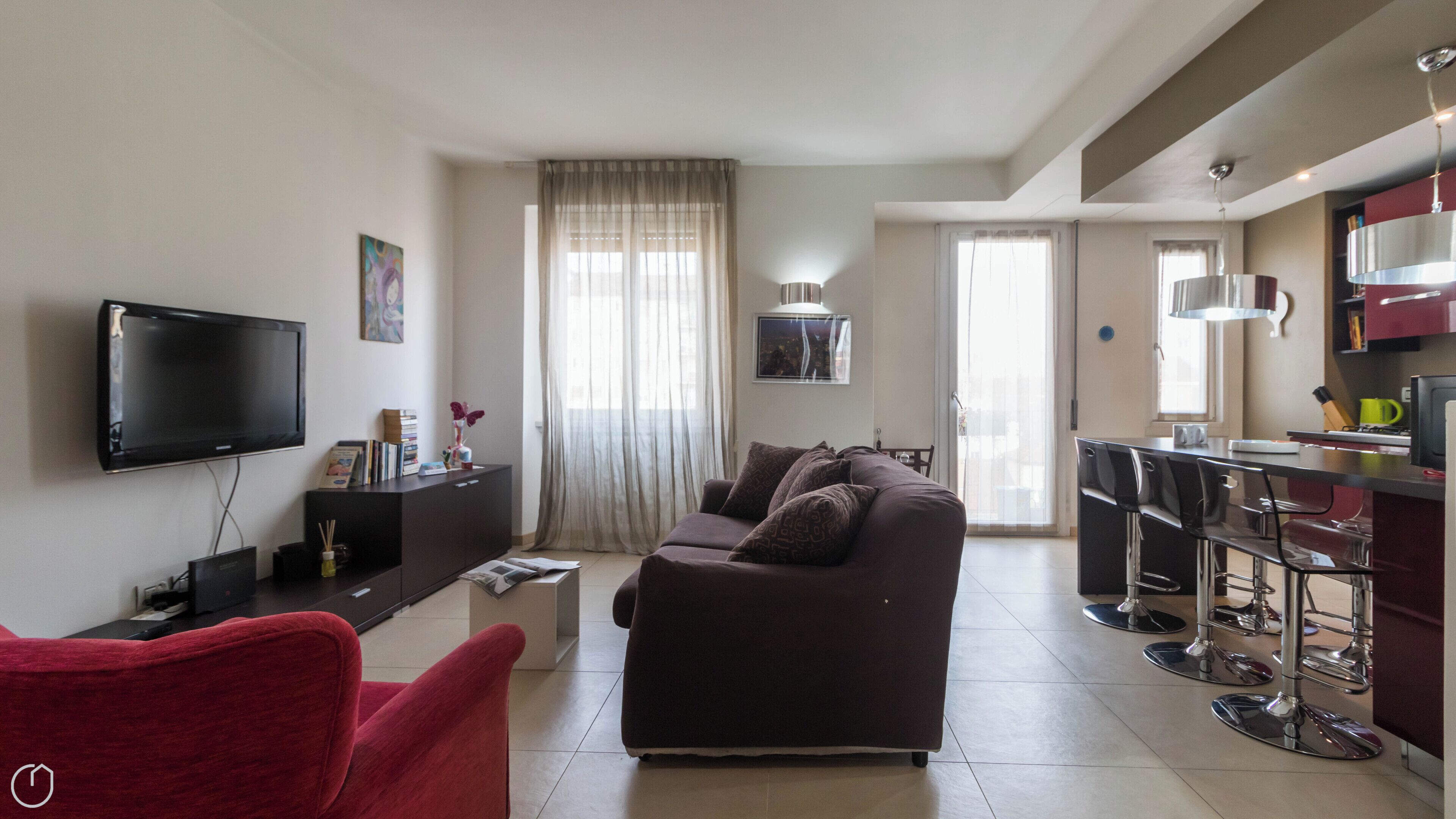 A modern living room with a flat-screen TV, a sofa, a red chair, and a kitchen area with bar stools.