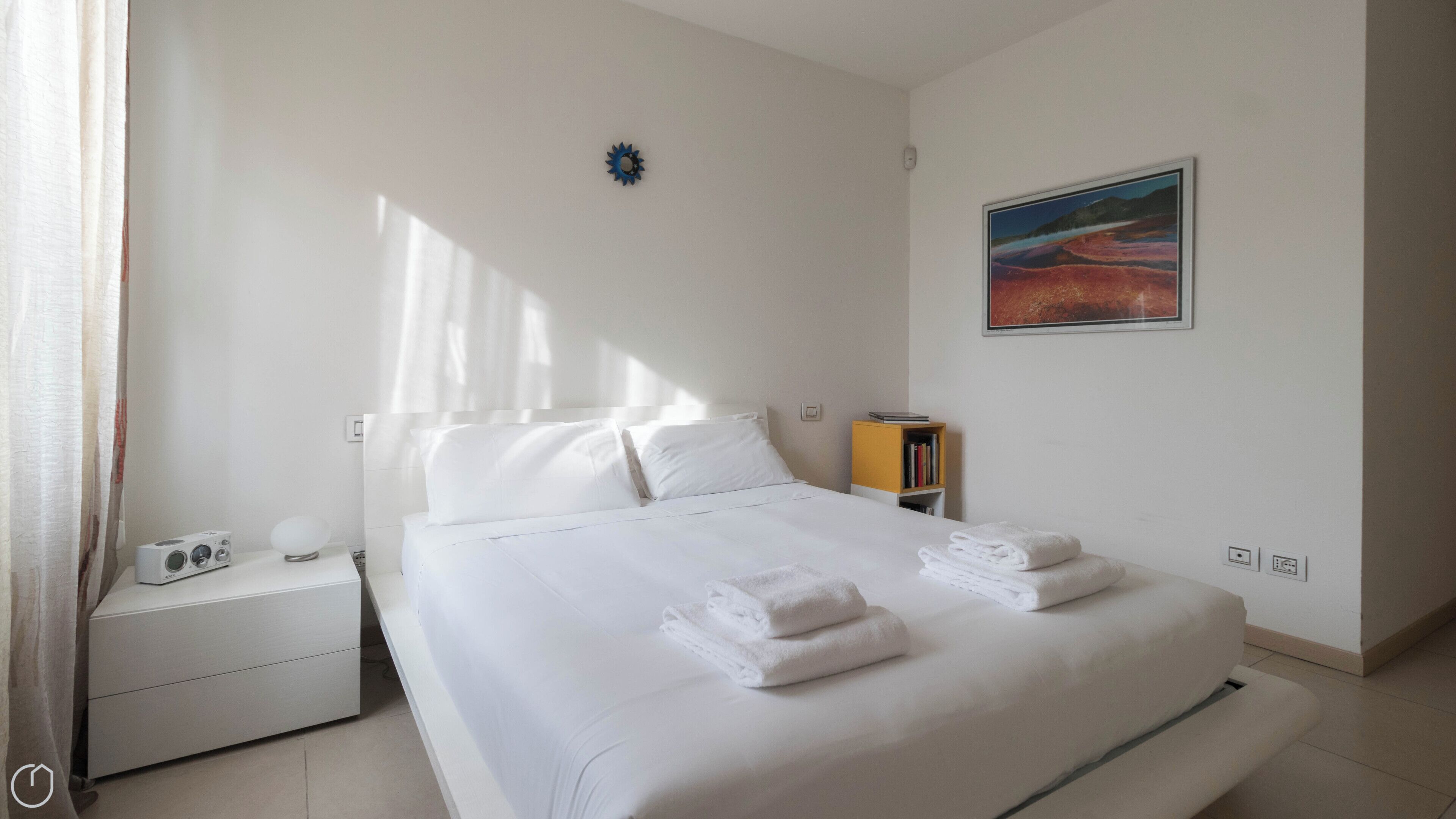 A neatly made bed with white linens and towels, a bedside table with a lamp and a small electronic device, a bookshelf with books, and a painting on the wall.