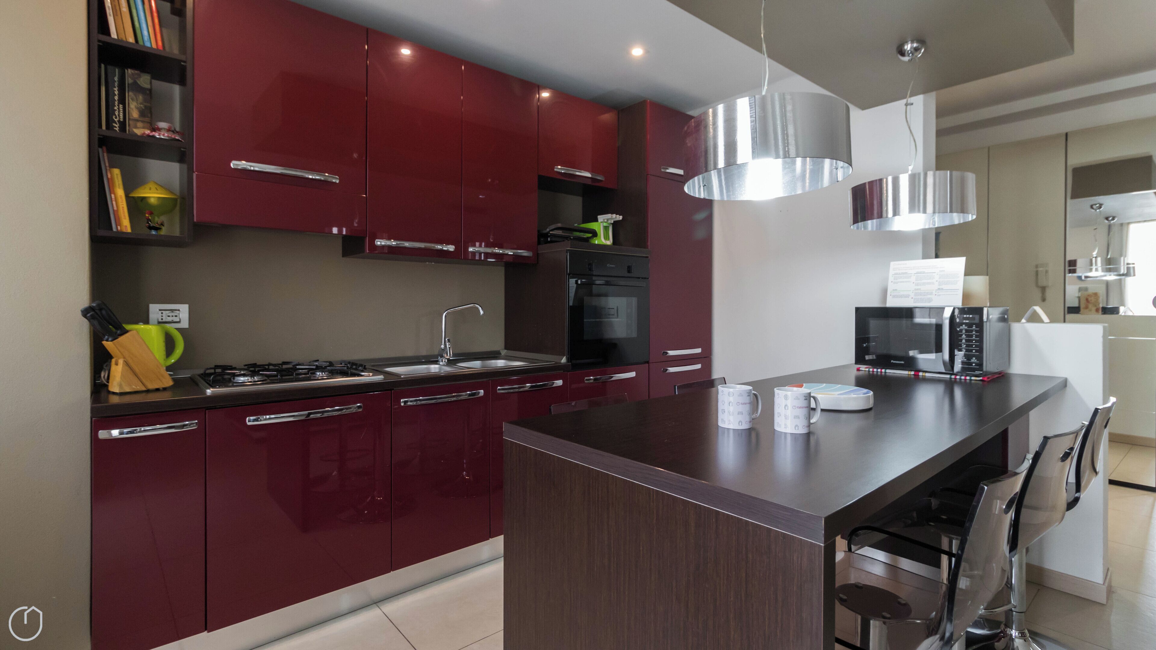 A modern kitchen with red cabinets, a central island, stainless steel appliances, and a dining area with chairs.