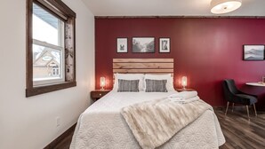 Une chambre avec un grand lit, une tête de lit en bois, une fenêtre et un mur rouge orné de tableaux encadrés.