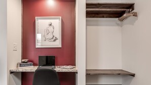 Une pièce avec un mur rouge, une œuvre d’art encadrée, un bureau avec un ordinateur portable et une chaise noire. Des étagères en bois se trouvent au-dessus du bureau.