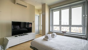 Una moderna camera d'albergo con un ampio letto, una televisione a schermo piatto, una finestra con vista sulla città e un comodino bianco.