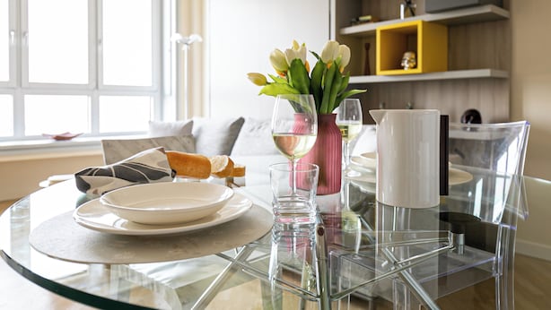 Un tavolo da pranzo con piano in vetro, un piatto bianco, un bicchiere di vino bianco, un vaso rosa con fiori e un panino.