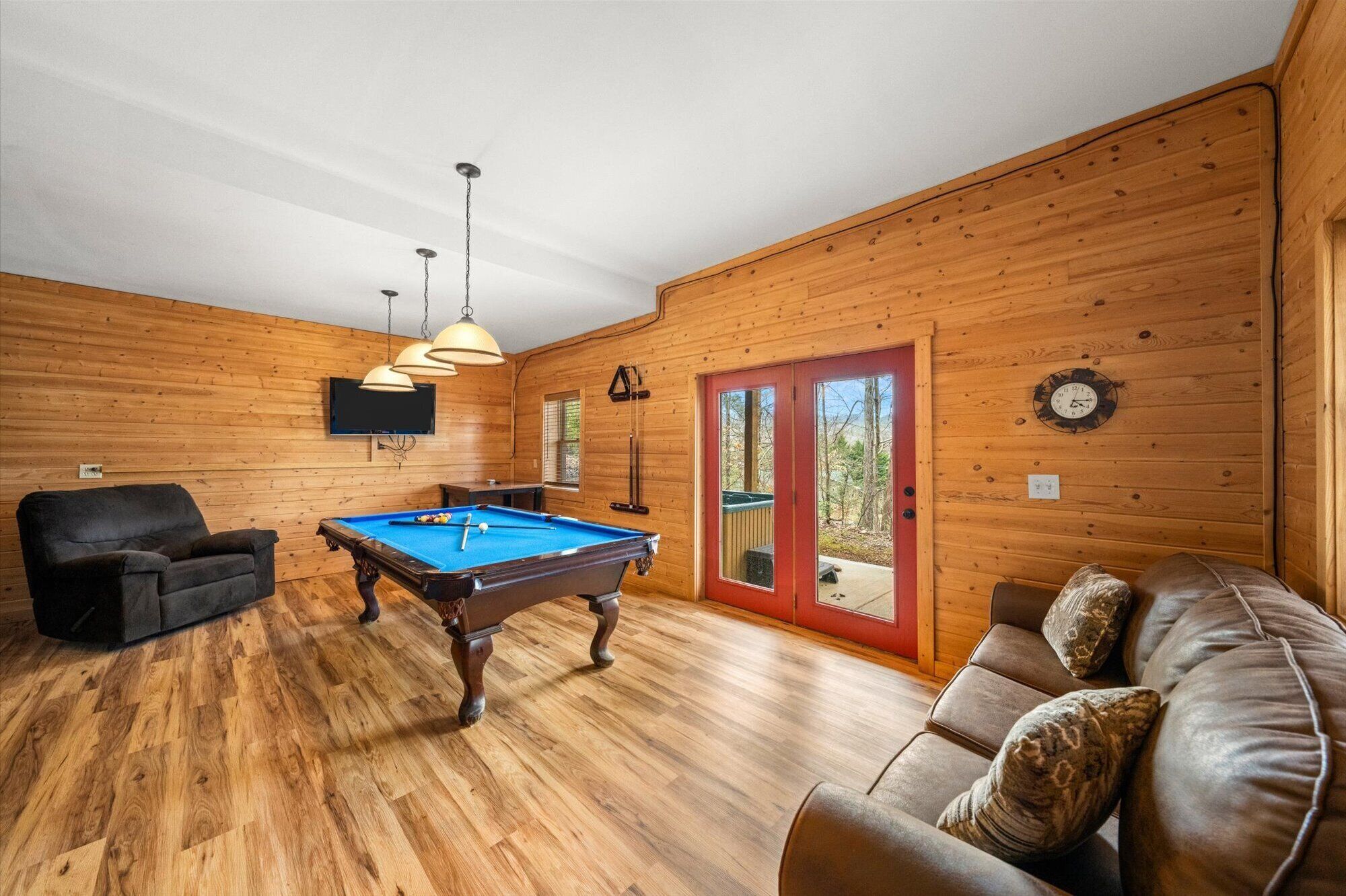 Een kamer met houten lambrisering, een pooltafel, een bank, een televisie en een deur naar buiten.