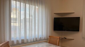 Een eetgedeelte met een tafel, stoelen, een aan de muur bevestigde televisie en dunne gordijnen.