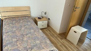 Een eenpersoonsbed met een gebedrukte spreidek, een nachtkastje met een lamp, een klein ladeblok en een houten deur met een handvat.