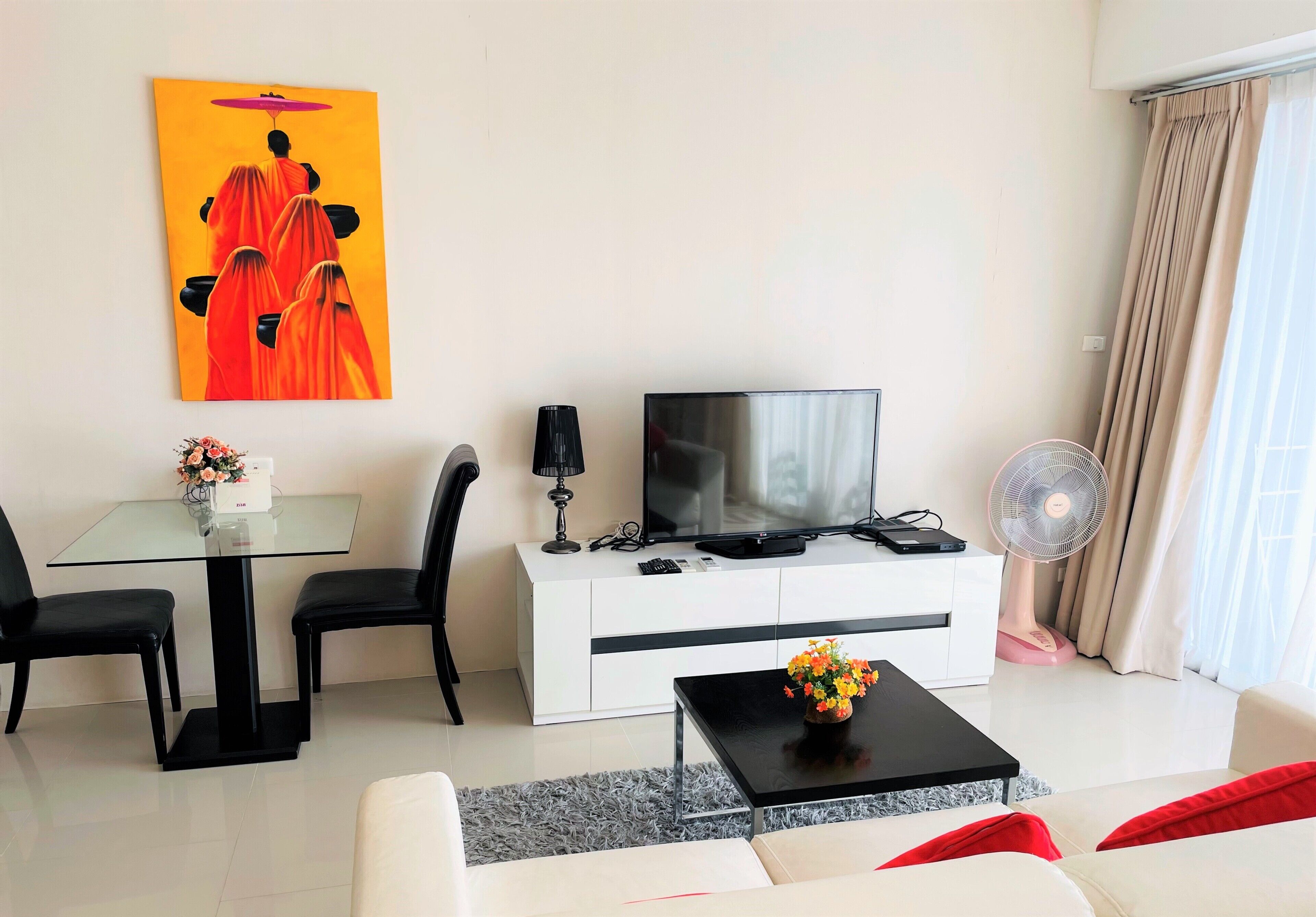 Een moderne woonkamer met een glazen tafel, zwarte stoelen, een bank met rode kussens, een televisie op een witte kast, een ventilator en een schilderij aan de muur.