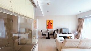 Quarto de hotel moderno com box de vidro, sofá branco, área de jantar com mesa e cadeiras, e uma TV em uma escrivaninha.