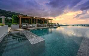 A serene infinity pool with a wooden pergola and lounge chairs, overlooking a lush green hillside and the ocean at sunset.