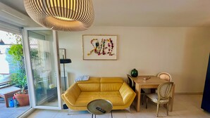 A modern living room with a yellow sofa, a glass coffee table, a dining table with chairs, a hanging light fixture, and a framed floral artwork on the wall. - Magnifique Design T2, Confortable, Parking Gratuit (Nice)